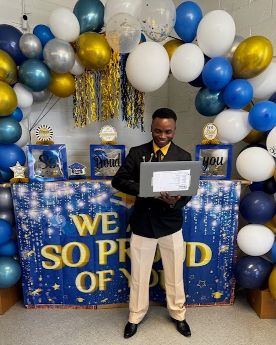 Jean Kiza holding a PCs for People Laptop in our Mission Market, with graduation decorations in the background.